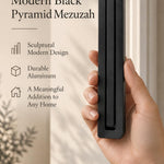 A hand presents the Large Modern Black Pyramid Mezuzah with its deep matte black aluminum mezuzah cover facing forward, highlighting the clean graduated bars and geometric vertical profile beside design illustrations for a contemporary Jewish doorway accent