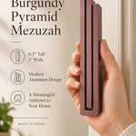 Hand holding the Large Modern Burgundy Pyramid Mezuzah upright, revealing its muted burgundy wine-red aluminum mezuzah cover, slender three-bar Pyramid design and graduated top beside illustrated product features for contemporary Jewish home Judaica