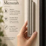 Large Modern Sandstone Pyramid Mezuzah held beside a doorpost, with the warm taupe sandstone aluminum mezuzah cover shown close enough to reveal its three stepped bars and rounded lower edge alongside product-name and design callouts for this contemporary Judaica piece