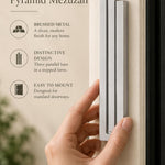 Large Modern Silver Pyramid Mezuzah held beside a doorpost, with the softly brushed silver aluminum mezuzah cover shown close enough to reveal its three stepped bars and rounded lower edge alongside product-name and design callouts for this contemporary Judaica piece