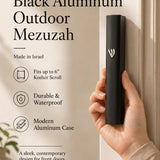 holding the large black aluminum outdoor mezuzah case beside a light doorframe, showing its silver Hebrew Shin and matte finish alongside text about its Israeli origin, six-inch kosher scroll fit and waterproof construction