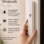 positioning the large modern white aluminum outdoor mezuzah case on a light doorpost beside a dark doorframe, showing its clean front face and raised Hebrew Shin