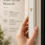 A hand holds the gold-framed white acrylic mezuzah with a prominent Hebrew Shin letter against a soft neutral background.