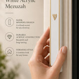 A hand holds the gold-framed white acrylic mezuzah with a prominent Hebrew Shin letter against a soft neutral background.