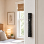 Modern Black Steps aluminum mezuzah mounted on a white bedroom doorpost, featuring a narrow architectural black profile, repeating horizontal fins, and a centered silver Shin in a warm contemporary interior