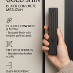 A hand holds a modern black concrete mezuzah case featuring a raised embossed Hebrew Shin with elegant gold trim.