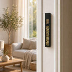 Modern Gold Crystal Inlay Black Cement Mezuzah mounted vertically on a residential doorpost, showing the slim matte black case, raised gold Shin, and shimmering golden crystal and glass inlay against a bright contemporary home interior