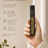Handheld modern black cement mezuzah case with a raised gold Shin and sparkling gold crystal and glass inlay, shown beside product information in a warm neutral setting to illustrate the case scale, dark cement finish, and decorative gold accents