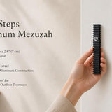 holding the Modern Black Steps aluminum mezuzah case beside a doorway, showing the slim layered black design and silver Shin, with Made in Israel construction, waterproof indoor and outdoor use, and fit for up to a 2.8-inch kosher mezuzah scroll