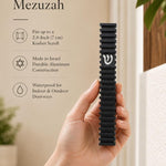 Hand holding the Modern Black Steps waterproof aluminum mezuzah case beside a doorway, showing its slim black stepped body and silver Shin; made in Israel and designed to fit up to a 2.8-inch kosher mezuzah scroll