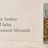 Modern gray cement mezuzah displayed upright in a minimalist setting with a raised silver-tone Shin and vertical golden amber crystal and glass inlay, highlighting warm honey, bronze, and gold reflections against the cool neutral cement surface
