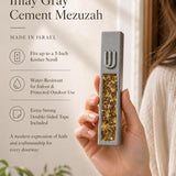 Handheld modern gray cement mezuzah case with a raised silver-tone Shin and golden amber crystal and glass inlay, presented beside product details in a soft neutral interior to show the slim case profile, natural-looking warm color variation, and reflective finish