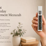 Hand holding a Modern Iridescent Crystal Inlay White Cement Mezuzah case with a raised silver-tone Shin and reflective crystal and glass inlay showing pearl, charcoal, bronze, blue-gray, green, and iridescent tones in a warm contemporary interior