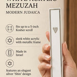 Close up of a hand holding a white and silver acrylic mezuzah with a prominent Shin letter in a bright editorial setting.