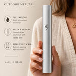 Premium handheld image of the modern waterproof silver aluminum mezuzah, held naturally with editorial text on the side, in soft neutral tones.