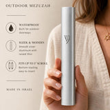 Premium handheld image of the modern waterproof silver aluminum mezuzah, held naturally with editorial text on the side, in soft neutral tones.