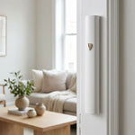 Modern white aluminum mezuzah case mounted on a white interior doorpost, with a small gold-tone Hebrew Shin on its curved face. A bright living room with a sofa and light wood table is visible through the doorway.