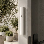 White aluminum outdoor mezuzah case mounted on a light-colored doorpost beside a dark entry door. Its narrow curved face carries a silver-tone Hebrew Shin, with a planted stone patio visible beyond the entrance.