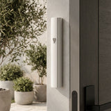 White aluminum outdoor mezuzah case mounted on a light-colored doorpost beside a dark entry door. Its narrow curved face carries a silver-tone Hebrew Shin, with a planted stone patio visible beyond the entrance.