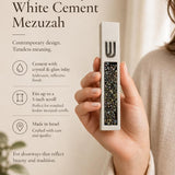 Handheld view of a modern white cement mezuzah case with a raised silver-tone Shin and dense iridescent crystal and glass inlay, shown beside product information in a neutral home setting to highlight the contemporary design, slim proportions, and reflective finish