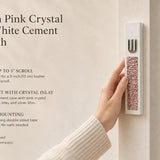 Handheld modern white cement mezuzah case featuring a raised silver-tone Shin and pink crystal and glass inlay, shown beside product information in a warm neutral home setting to highlight its slim profile, soft rose color palette, and contemporary styling
