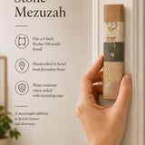A person holding a natural Jerusalem stone mezuzah case with gold Hebrew lettering, captured in warm, soft natural light.