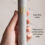 Person holding a gray cement Mezuzah with gold highlights, with curvy arrow annotations pointing to features, in a light background.