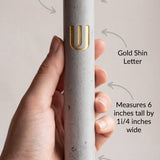 Person holding a gray cement Mezuzah with gold highlights, with curvy arrow annotations pointing to features, in a light background.
