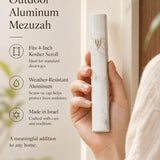 Person holding sleek white marble-inspired outdoor aluminum mezuzah with Hebrew Shin and gold accents.