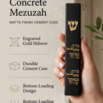 Close up of a hand holding an Israeli made black cement mezuzah case with an engraved gold Shin and Hebrew blessing.
