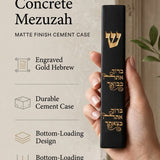 Close up of a hand holding an Israeli made black cement mezuzah case with an engraved gold Shin and Hebrew blessing.