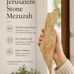 Editorial close-up of a hand holding an authentic Jerusalem stone mezuzah case with raised gold Land of Israel artwork.