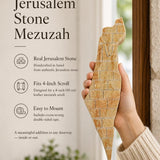 Editorial close-up of a hand holding an authentic Jerusalem stone mezuzah case with raised gold Land of Israel artwork.