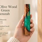 Hand holding a 7.5-inch olive wood and emerald green resin mezuzah featuring a gold-toned Shin at a Jewish entrance.