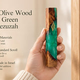 Hand holding a 7.5-inch olive wood and emerald green resin mezuzah featuring a gold-toned Shin at a Jewish entrance.