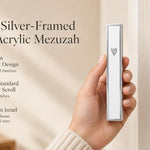 A hand holding a modern silver-framed white acrylic mezuzah with a silver Shin against a soft neutral background.