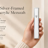 A hand holding a modern silver-framed white acrylic mezuzah with a silver Shin against a soft neutral background.
