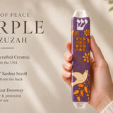 Hand-holding purple Dove of Peace mezuzah with white dove motif and orange accents, placed beside editorial text for premium presentation.