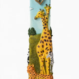 Alternate front view of the Savannah Safari Fimo Mezuzah mezuzah cover, showing a pale blue Fimo body with tall yellow giraffes, a zebra, spotted animals, green scenery and a gold-colored Hebrew Shin beneath decorative brass fittings and the full decorative shape in a closer product photograph of this distinctive Jewish doorway design