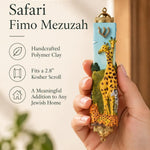Handheld portrait of the Savannah Safari Fimo Mezuzah mezuzah cover, with yellow giraffes, a zebra and spotted animals against a pale blue Fimo body, with green scenery, a gold-colored Shin and decorative brass fittings clearly visible beside the product name and illustrated features of this distinctive Jewish doorway design