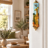 Savannah Safari Fimo children’s mezuzah cover mounted vertically on a pale interior doorpost, with yellow giraffes, a zebra and spotted animals rising from green scenery beneath a gold-colored Hebrew Shin on the pale blue body