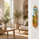 Savannah Safari Fimo Mezuzah mounted vertically on a pale home doorpost, with a pale blue Fimo body with tall yellow giraffes, a zebra, spotted animals, green scenery and a gold-colored Hebrew Shin beneath decorative brass fittings creating a distinctive mezuzah cover that brings its Jewish design and decorative detail into the doorway