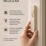 hand holds the Small Modern Cream Jerusalem Stone Mezuzah beside a light doorpost, showing its narrow stone body and gold Shin alongside graphic callouts about the natural stone and scroll chamber