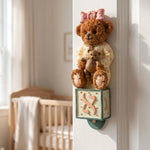 Hand-painted Teddy Aleph Bear Mezuzah case in soft cream, pink, brown, and green tones, mounted inside a doorway in a nursery or child's room.