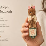 Hand holding Teddy Aleph Bear Mezuzah with soft nursery colors and Hebrew letter Aleph for a child's room decor.