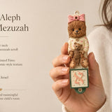 Hand holding Teddy Aleph Bear Mezuzah with soft nursery colors and Hebrew letter Aleph for a child's room decor.