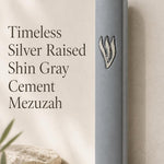 Timeless gray cement mezuzah displayed upright against a minimalist neutral background, featuring a large raised sculptural silver-tone Shin on the smooth gray face and a clean understated contemporary Judaica design