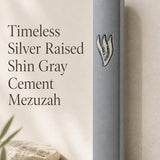 Timeless gray cement mezuzah displayed upright against a minimalist neutral background, featuring a large raised sculptural silver-tone Shin on the smooth gray face and a clean understated contemporary Judaica design
