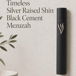 Close product portrait of the Timeless Black Cement Mezuzah featuring a prominent raised silver-tone Shin against the smooth dark surface, emphasizing the sculptural Hebrew letter, monochrome black case, and understated modern design