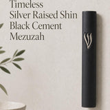 Close product portrait of the Timeless Black Cement Mezuzah featuring a prominent raised silver-tone Shin against the smooth dark surface, emphasizing the sculptural Hebrew letter, monochrome black case, and understated modern design